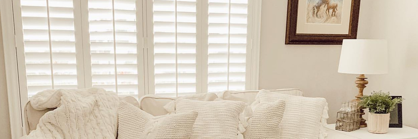 White and beige living room with white polywood shutters above a white couch.