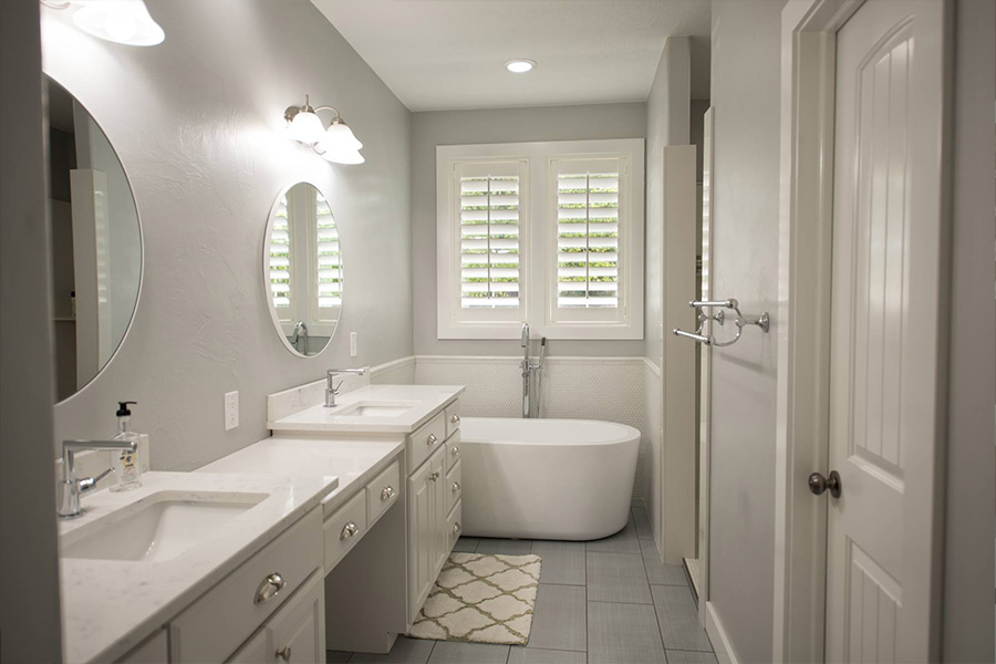 White and gray bathroom with one white Polywood shutters on a window above a modern style bathtub.